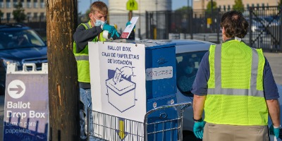 Image of the Board of Elections official ballot drop box, including two men in bright vests accepting ballots from passing cars